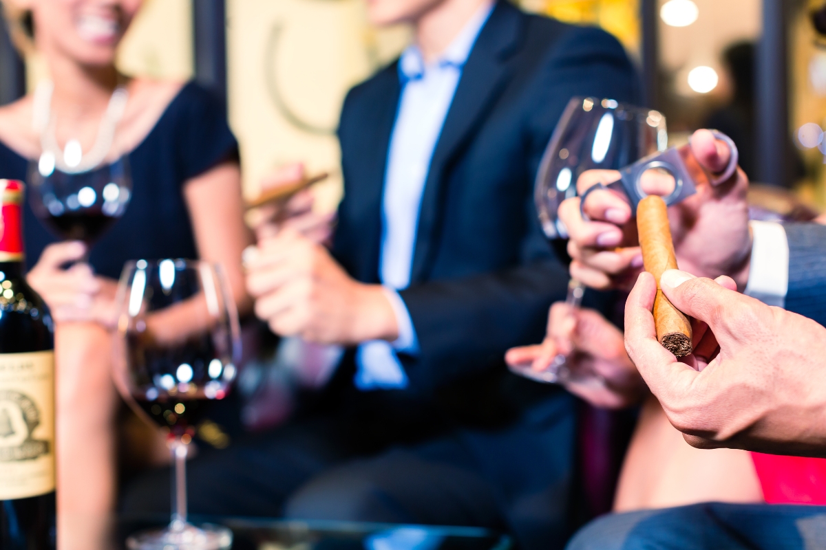 Person holding a cigar with wine glasses in the background, at a cigar club gathering.