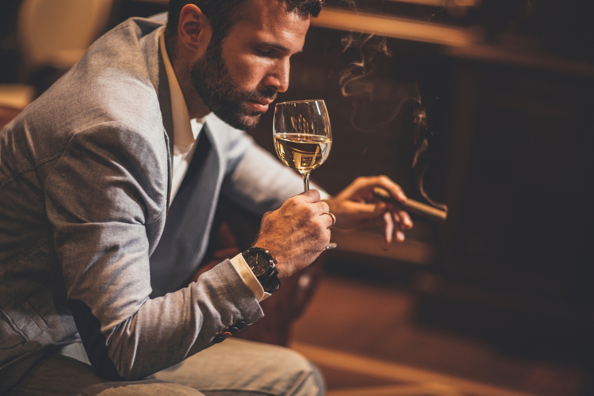 A well-dressed man holding a cigar responsibly in one hand and a glass of white wine in the other, sitting pensively in an elegantly furnished room.