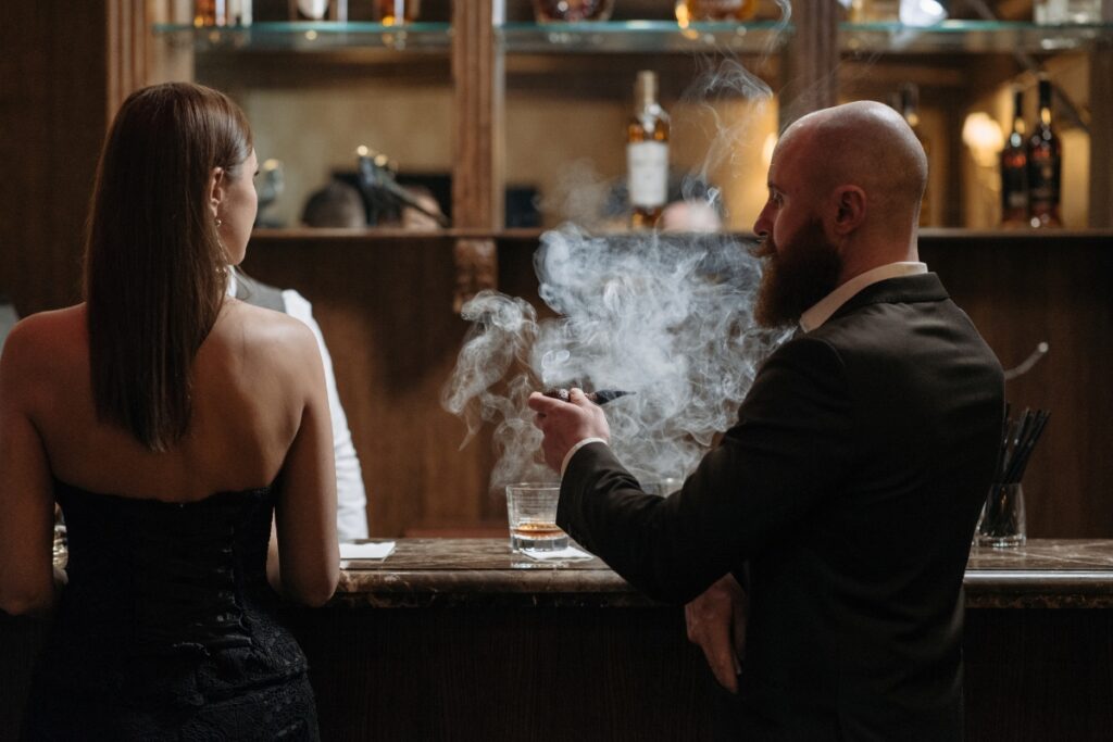 A bald man in a suit smoking a pipe, sitting at a bar next to a woman in a black dress, with drinks on the counter.