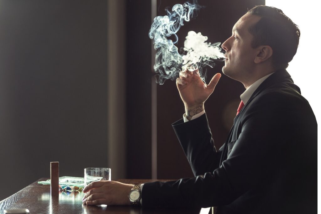 A man in a suit exhales smoke from a cigar while sitting at a table with a glass of whiskey, evoking a contemplative atmosphere.
