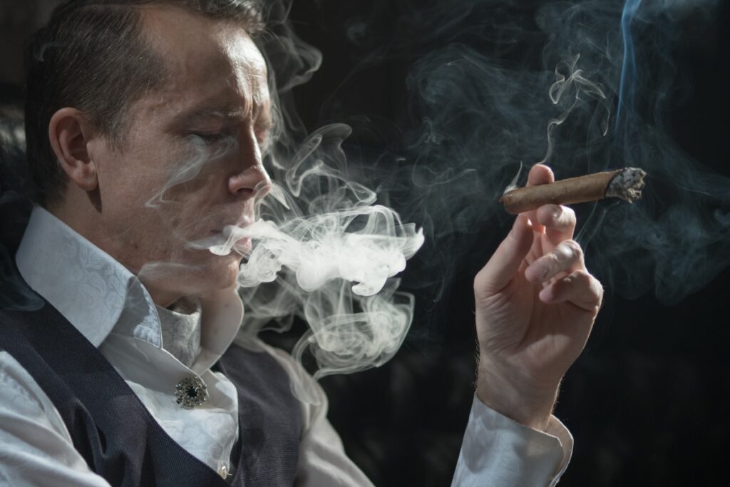 A man in a formal vest exhales smoke while holding a cigar at a cigar and pipe event, with smoke swirling around him in a dimly lit setting.