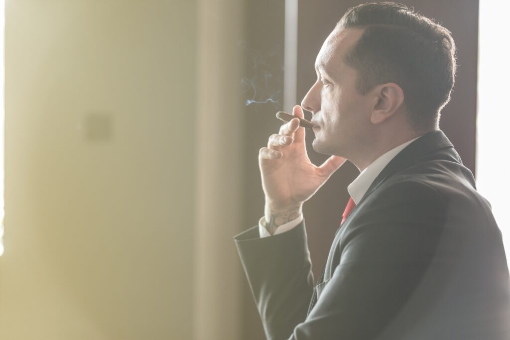 A man in a suit smoking a cigar indoors, bathed in soft sunlight from the window beside him.