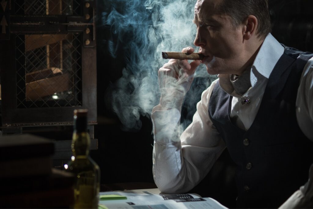 A man in a vest and tie exhaling smoke from a pipe, sitting by a table with a bottle and a document, in a dimly lit room.