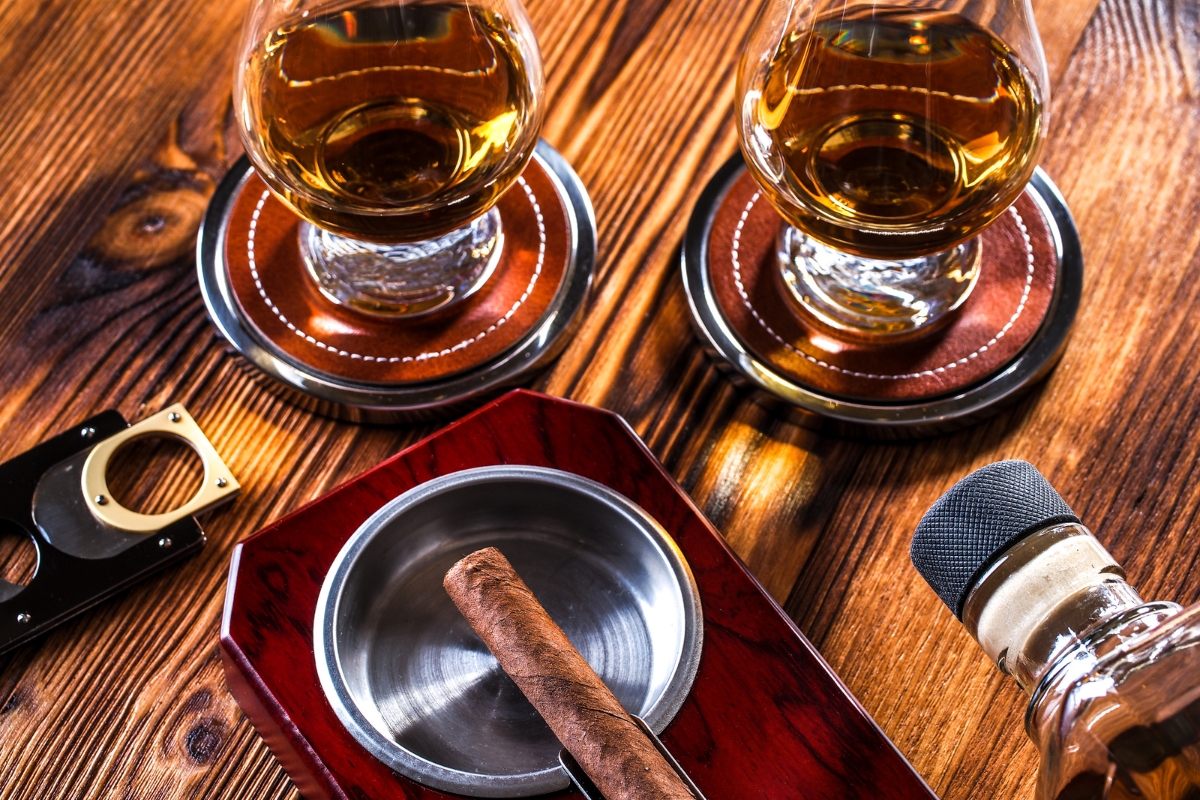 Two glasses of whiskey on wooden coasters, a famous cigar aficionado's cigar in an ashtray, a cigar cutter, and a bottle, on a wooden table.