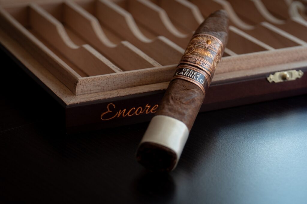 A single cigar, favored by famous cigar aficionados, rests on a dark wooden table, partially on a cigar box labeled "encore" by Perez Carrillo.