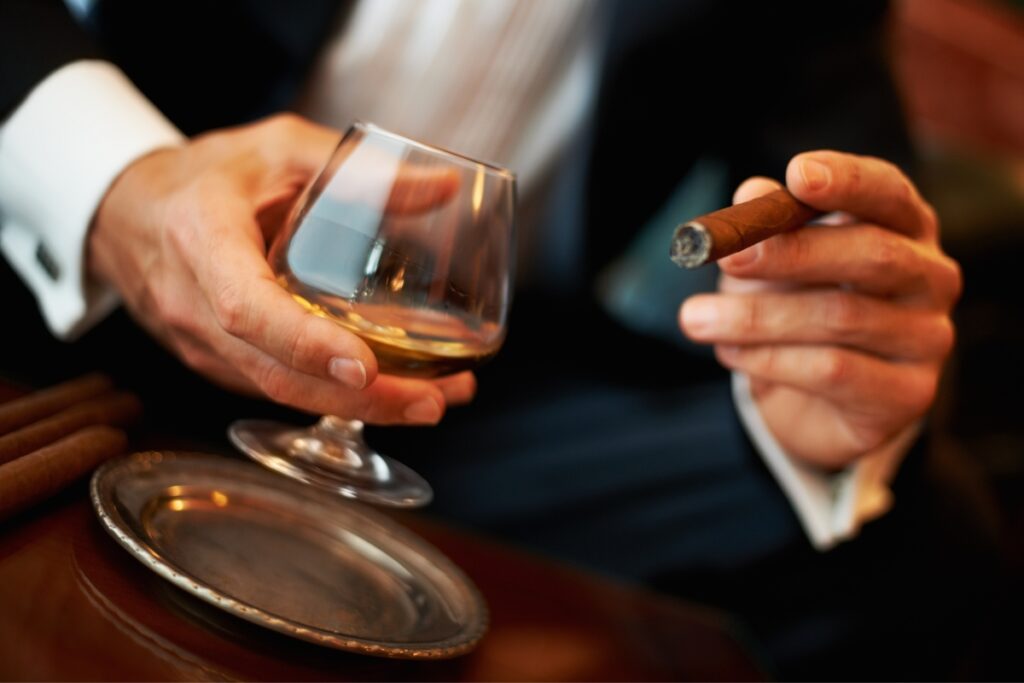 A well-dressed man, known among famous cigar aficionados, holds a glass of whiskey in one hand and a lit cigar in the other, with cigars on a silver tray in the foreground.