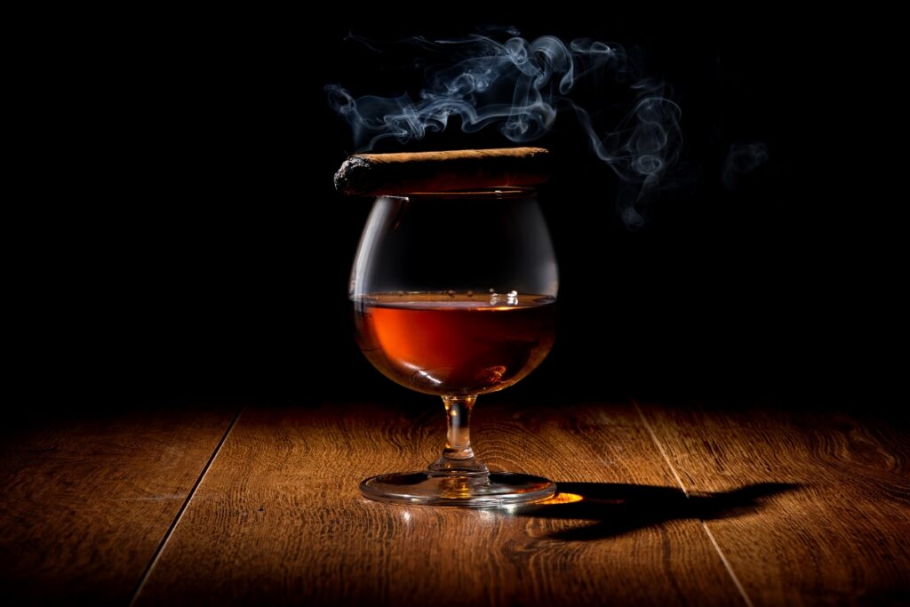 A snifter of amber-colored liquor with a smoking cigar resting on top, placed on a wooden table, highlighted by a dark background, attracting famous cigar aficionados.