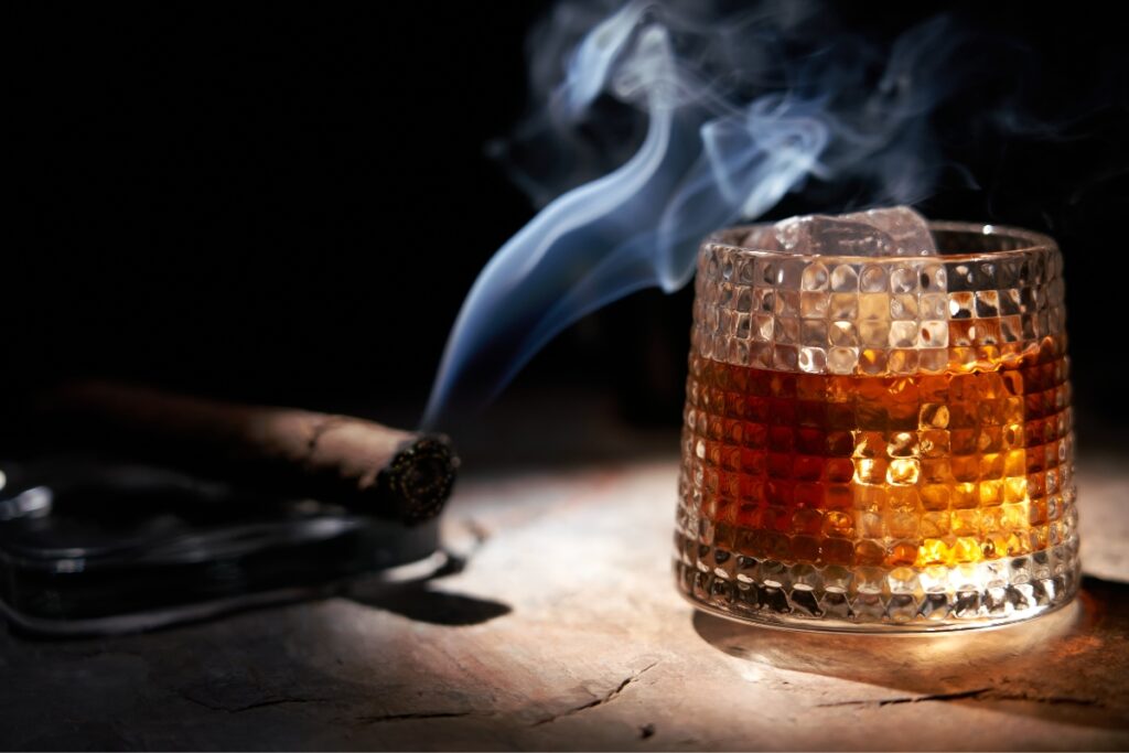 A glass of whiskey with ice next to a lit cigar emitting smoke, ideal for famous cigar aficionados, on a dark background.