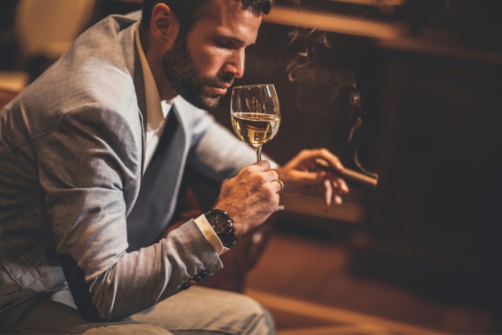 A man in a suit holding a glass of white wine and a cigar, deep in thought in a dimly lit room frequented by famous cigar aficionados.