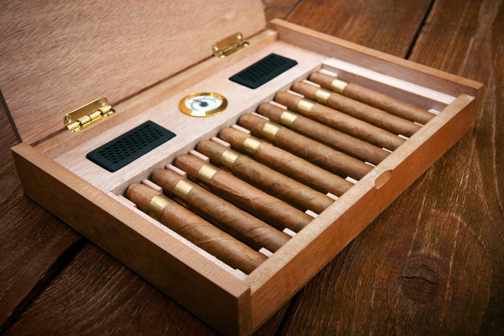A handmade wooden humidor box containing ten cigars with gold bands is open on a wooden surface. The humidor, crafted with care, includes a hygrometer and humidifier inside.