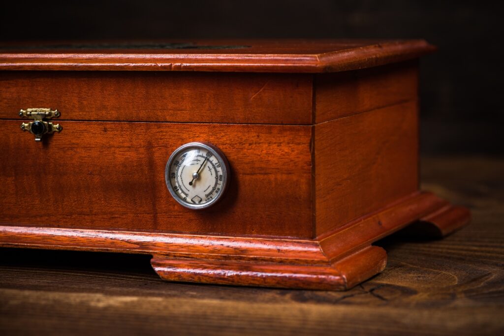A handmade humidor crafted from fine wood features a circular temperature gauge and a small latch on the front, resting elegantly on the wooden surface.