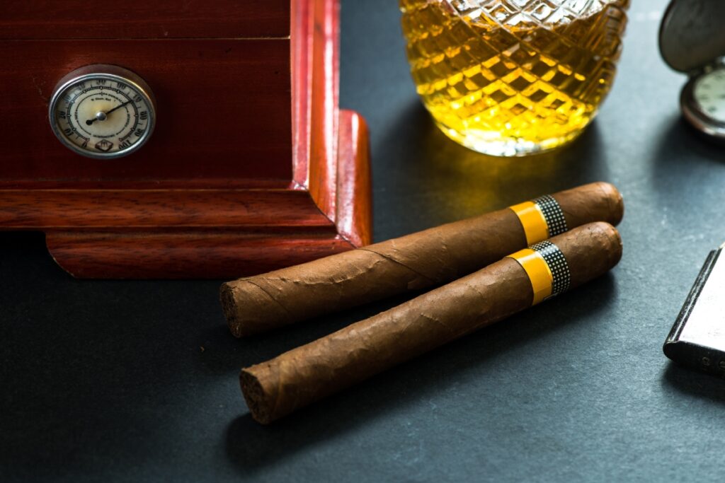 Two cigars, a wicker glass container with a yellow liquid, and part of a thermometer are placed on a dark surface. Nearby, handmade humidors add an element of crafted elegance to the scene.