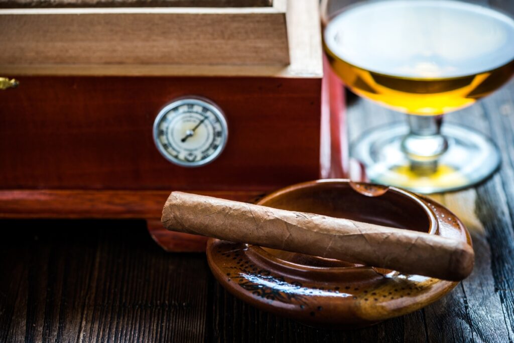 A cigar rests in an ashtray next to a glass of amber-colored liquid and a wooden box with a hygrometer, showcasing the elegance of handmade humidors.