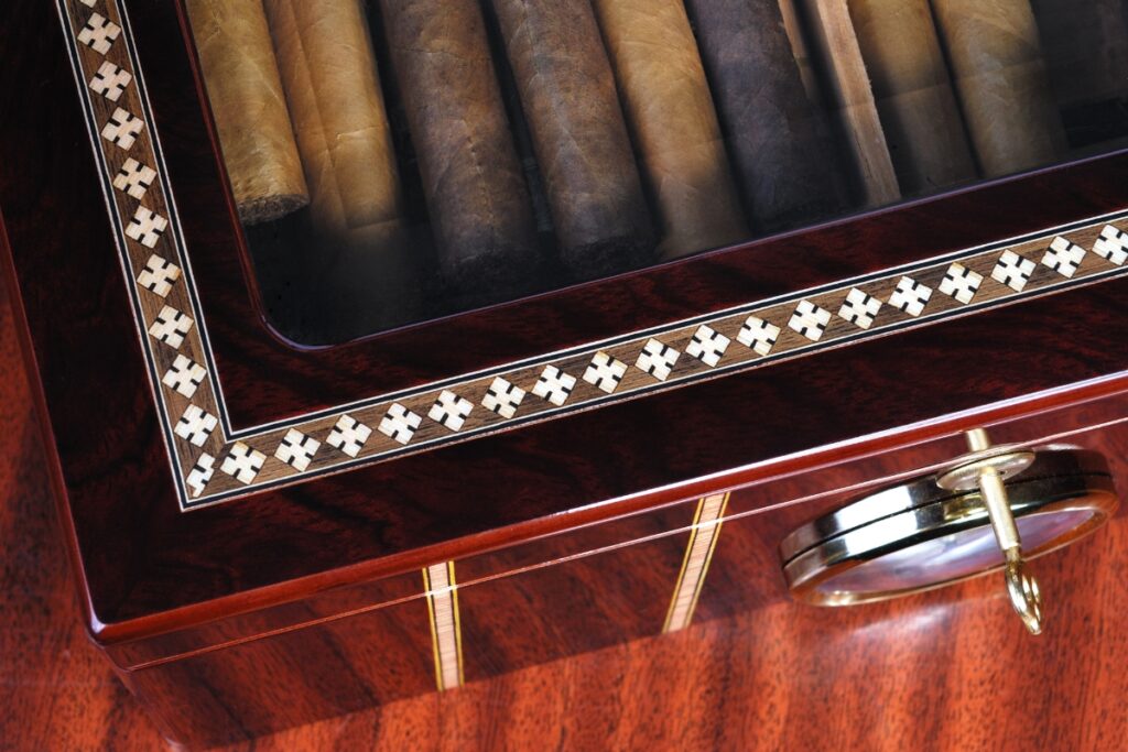 A handmade humidor with a decorative border containing cigars visible through a glass lid, resting on a wooden surface.