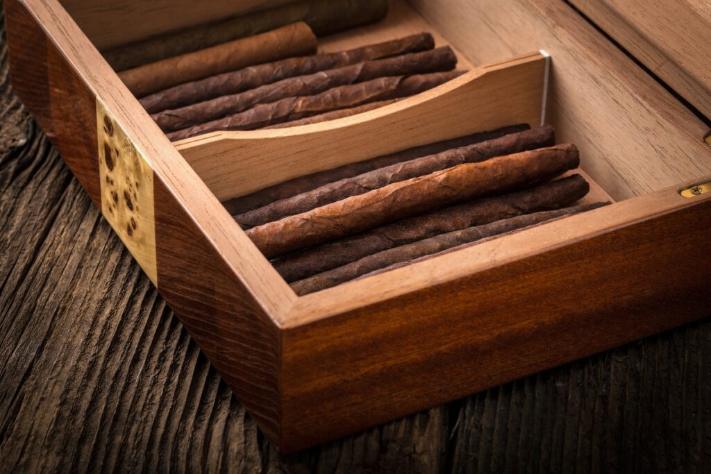 A handmade humidor with several cigars placed inside, partially divided by a wooden separator, rests on a wooden surface.