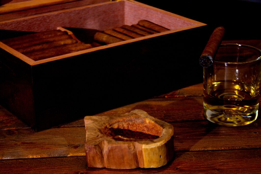 A wooden box of cigars, an ashtray, and a glass of whiskey rest on a dark wooden table beside exquisite handmade humidors.