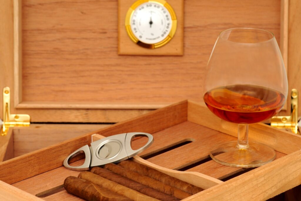 A handmade humidor containing cigars, a cigar cutter, and a glass of amber liquid, possibly cognac, with a hygrometer in the background.