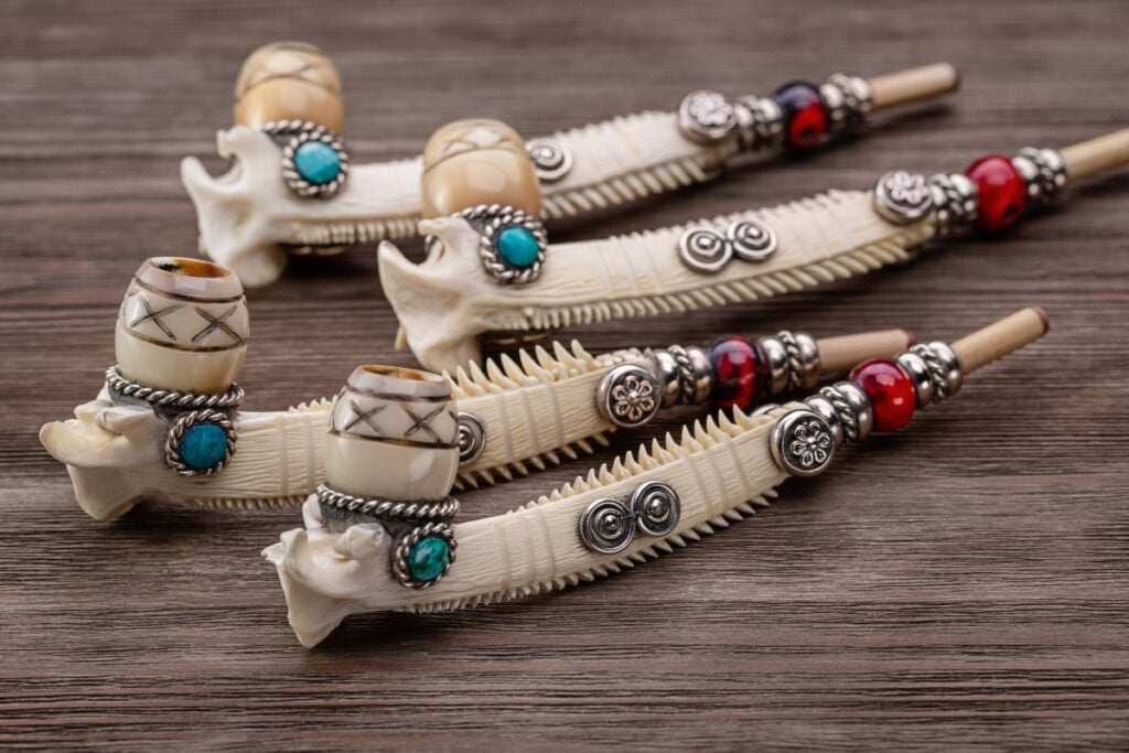 Carved bone smoking pipes with intricate designs and metal ornaments, including turquoise and red beads, reflecting Native American tobacco history, are displayed on a wooden surface.