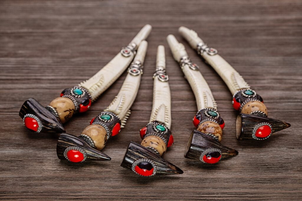 A set of five intricately designed ceremonial knives with carved handles, red gem accents, and ornate metal fittings, reminiscent of Native American craftsmanship, laid out on a wooden surface, evoking a deep sense of history.