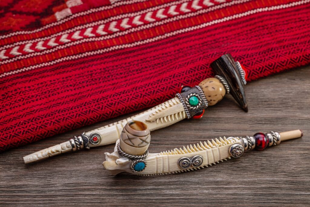 Two intricately decorated smoking pipes made from carved bone, adorned with turquoise and silver accents, placed on a wooden surface with a red patterned fabric in the background, evoke the rich tapestry of Native American tobacco history.