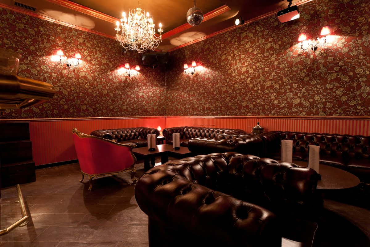 An elegant, dimly lit home smoking lounge with dark leather sofas, a red-patterned wall, and a chandelier hanging from the ceiling. A pink armchair and several small tables are also present.