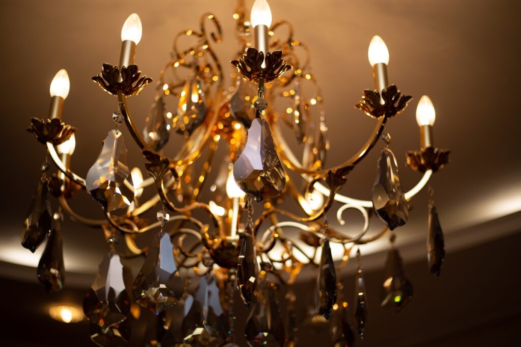 A close-up view of an ornate, gold-colored chandelier with crystal pendants, lit with several candle-style bulbs against a softly illuminated ceiling in an elegant home smoking lounge.