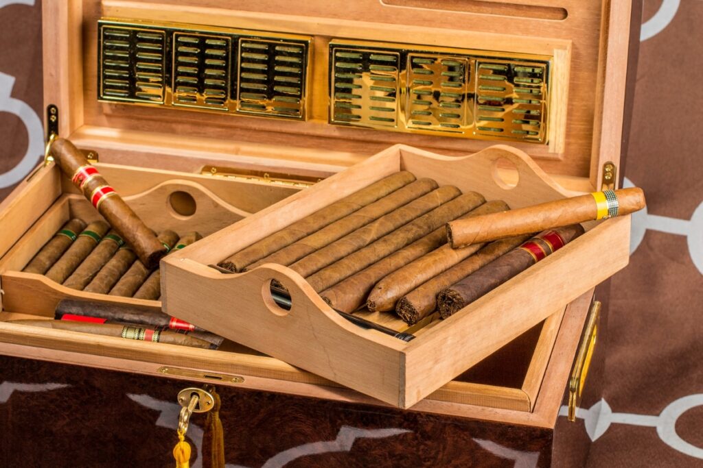 A wooden humidor box perfect for your home smoking lounge, containing various cigars in two compartments, with some cigars elegantly displayed on a tray placed atop the open box.