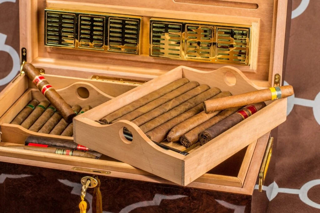 A wooden humidor box perfect for your home smoking lounge, containing various cigars in two compartments, with some cigars elegantly displayed on a tray placed atop the open box.