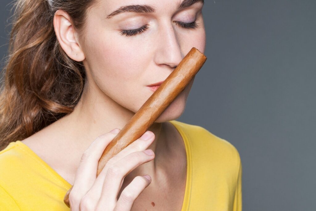 A woman in a yellow top is holding a large luxury cigar close to her nose with her eyes closed.