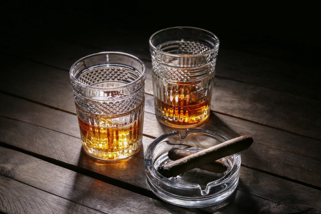 Two crystal glasses filled with amber liquid sit on a wooden table next to a glass ashtray holding two luxury cigars.