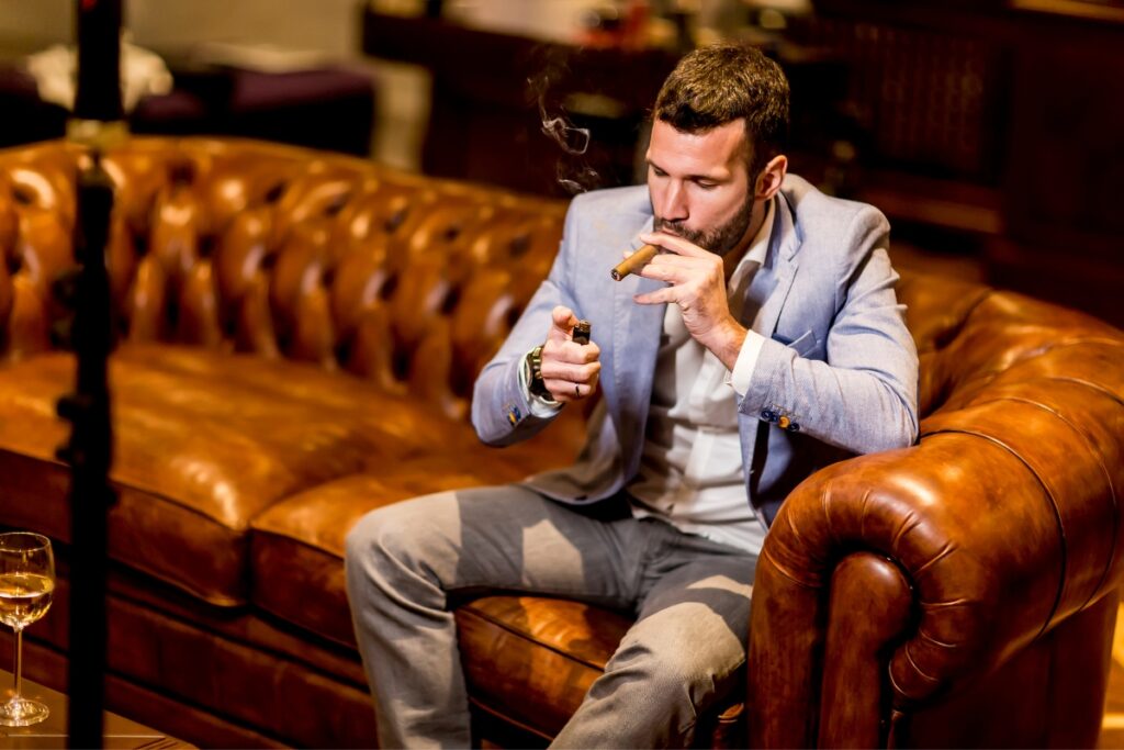 A man in a light blue blazer and grey pants sits on a brown leather sofa, lighting a luxury cigar with a lighter. A glass with a drink is on the table nearby.