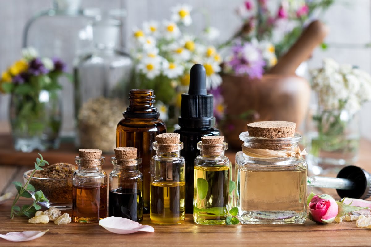 Assorted essential oil bottles with dropper and cork tops are arranged on a wooden surface, surrounded by fresh flowers and herbal plants. Perfectly selected, these oils can be especially beneficial as an essential oil for smokers seeking a natural remedy.