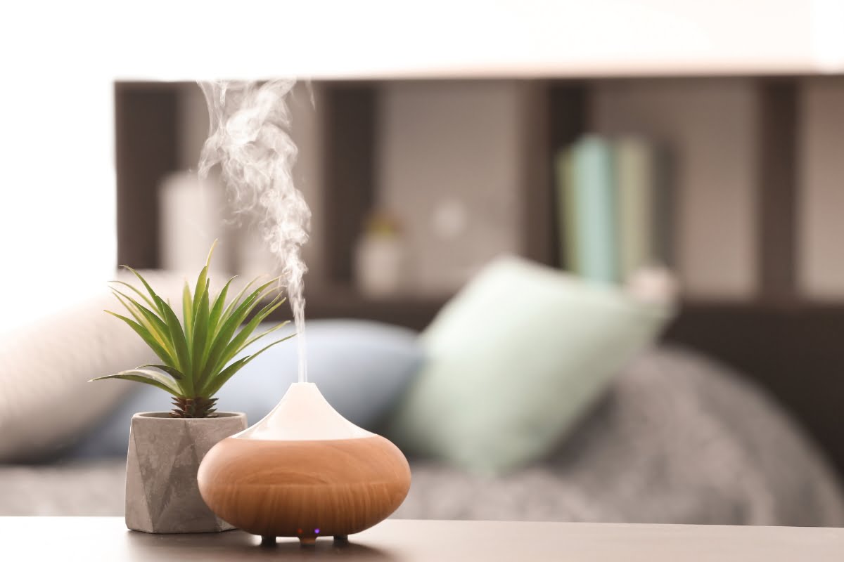 A wooden essential oil diffuser emits vapor on a table next to a small potted plant in a cozy bedroom, infusing the air with an essential oil for smokers, creating a serene and refreshing atmosphere.