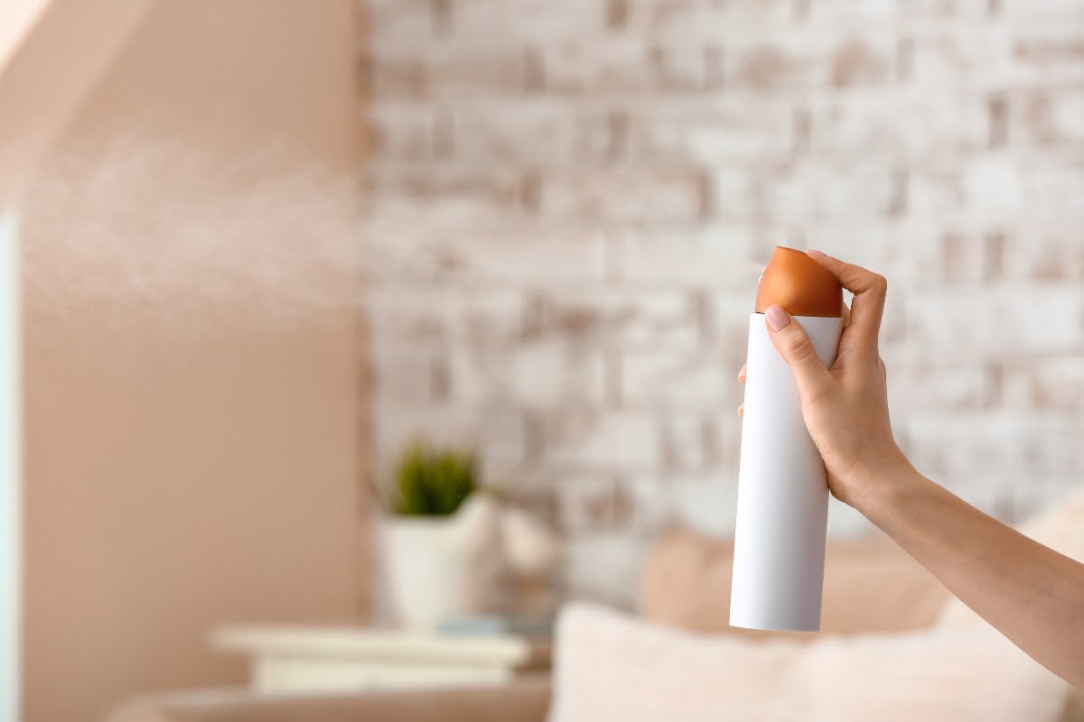 A hand holding and spraying an air freshener with essential oil for smokers in a room with a blurred, brick wall background.