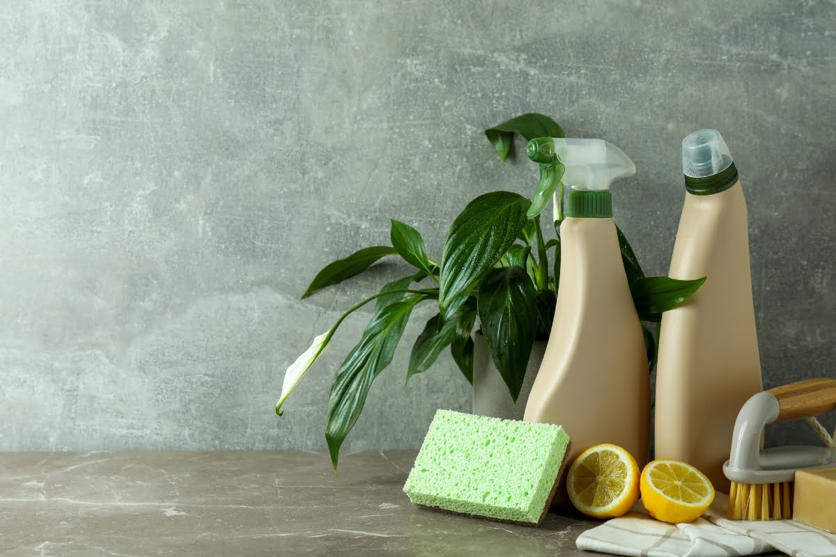 Cleaning supplies including spray bottles, a sponge, a scrub brush, lemon halves, two towels, and a green plant next to a gray wall. An essential oil for smokers is also included to help freshen the air.