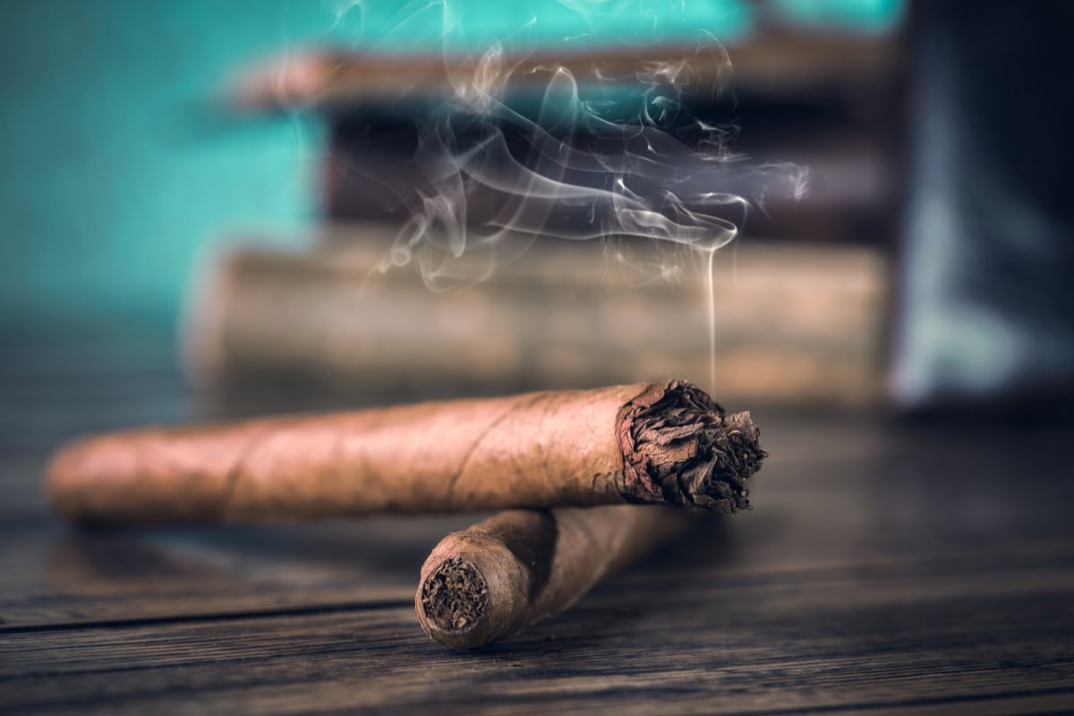 Two cigars on a wooden surface, one of which is lit and producing smoke, evoke a rich, aromatic ambiance. In the blurred background, the subtle scent of essential oil for smokers adds an unexpected layer to the scene.