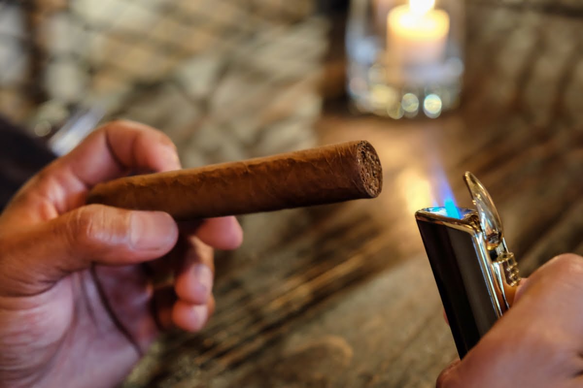 A person lighting a cigar with a blue-flame lighter on a wooden table, reminiscent of historical cigar smokers, with a lit candle glowing softly in the background.