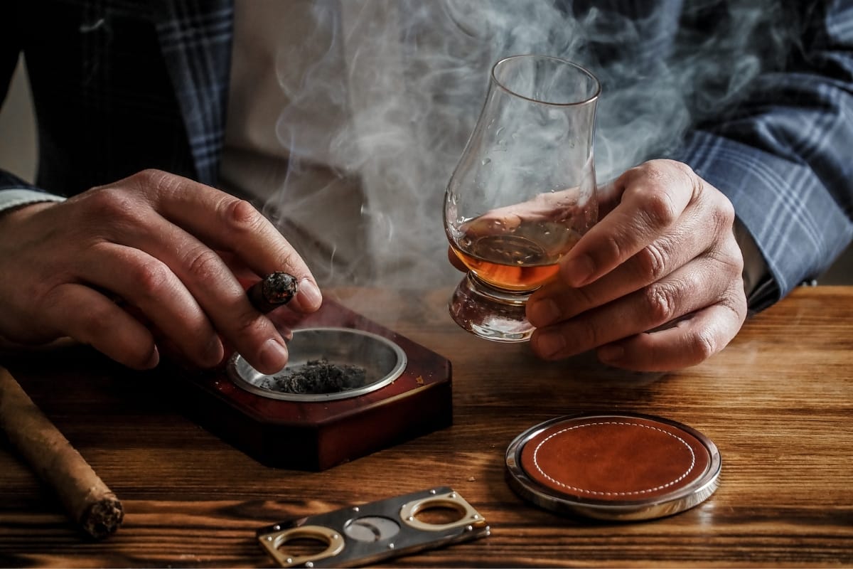 Person holding a glass of amber liquid in one hand and a lit cigar in the other, with a cigar cutter and ashtray on a wooden table, setting the scene for an exquisite food pairing experience.