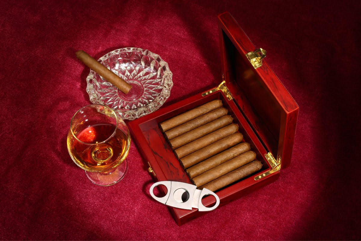 A wooden box of cigars, a cigar cutter, a glass of whiskey, and a lit cigar in an ashtray are set on a red velvet surface, creating the perfect display of smoking memorabilia.