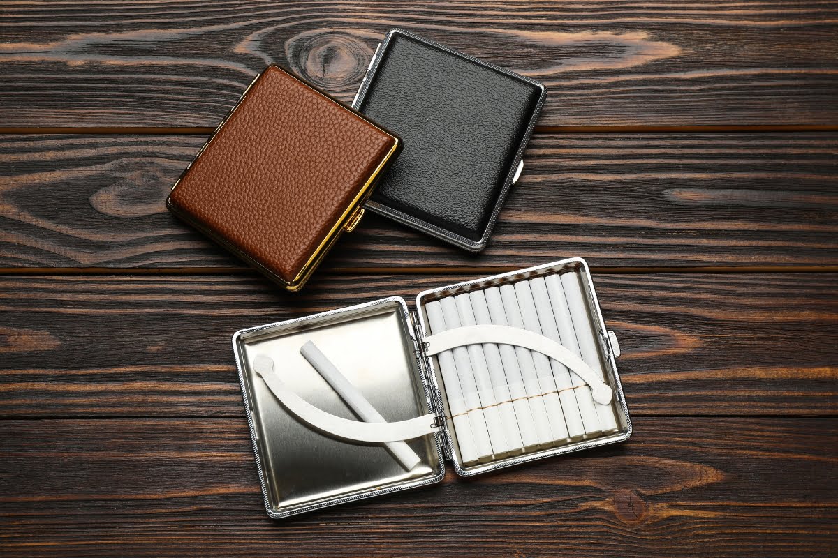 Two cigarette cases; one closed with a brown textured cover, another closed with a black cover, and an open metal case showing cigarettes on a wooden surface—perfect smoking memorabilia for any collector.