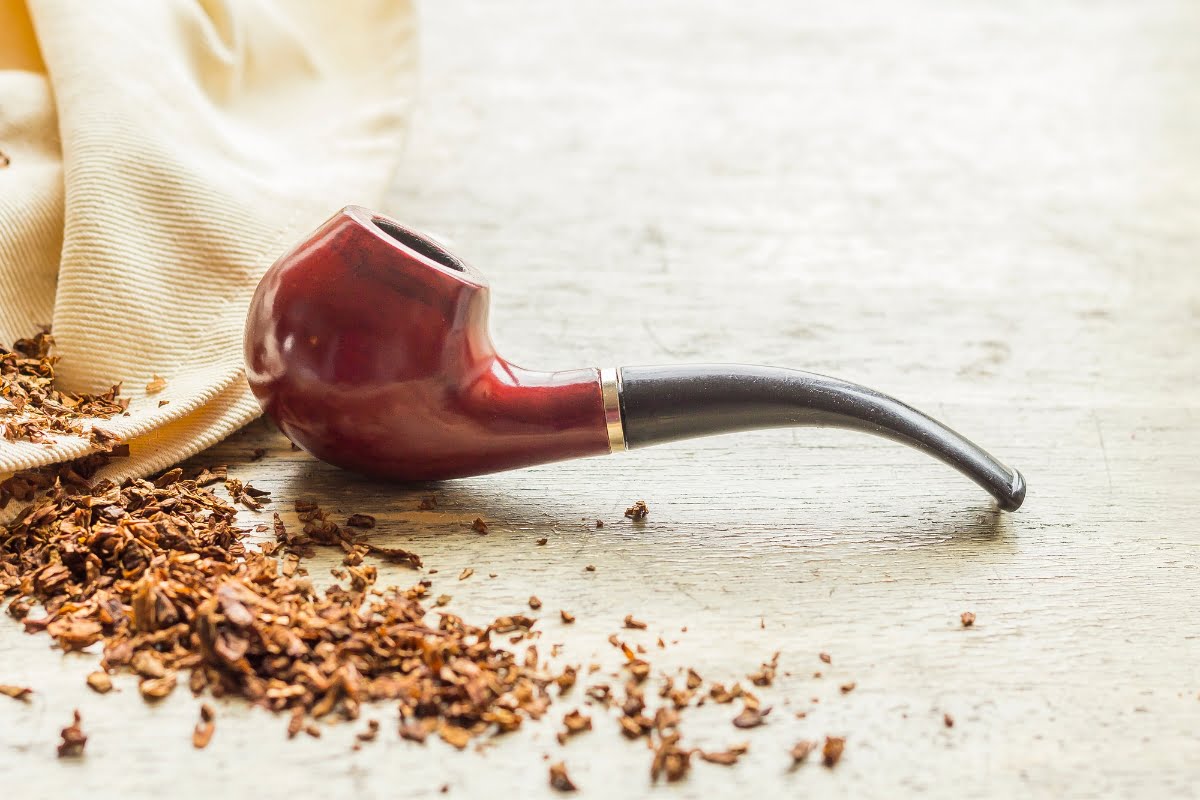 A piece of smoking memorabilia, this wooden tobacco pipe with a curved stem rests beside loose tobacco and a beige cloth on a wooden surface.