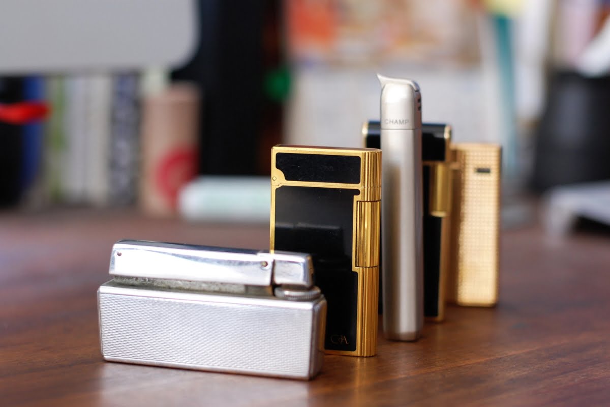 Close-up of various metal lighters, iconic smoking memorabilia, arranged in a row on a wooden surface. They feature gold, silver, and black designs with a blurred background.