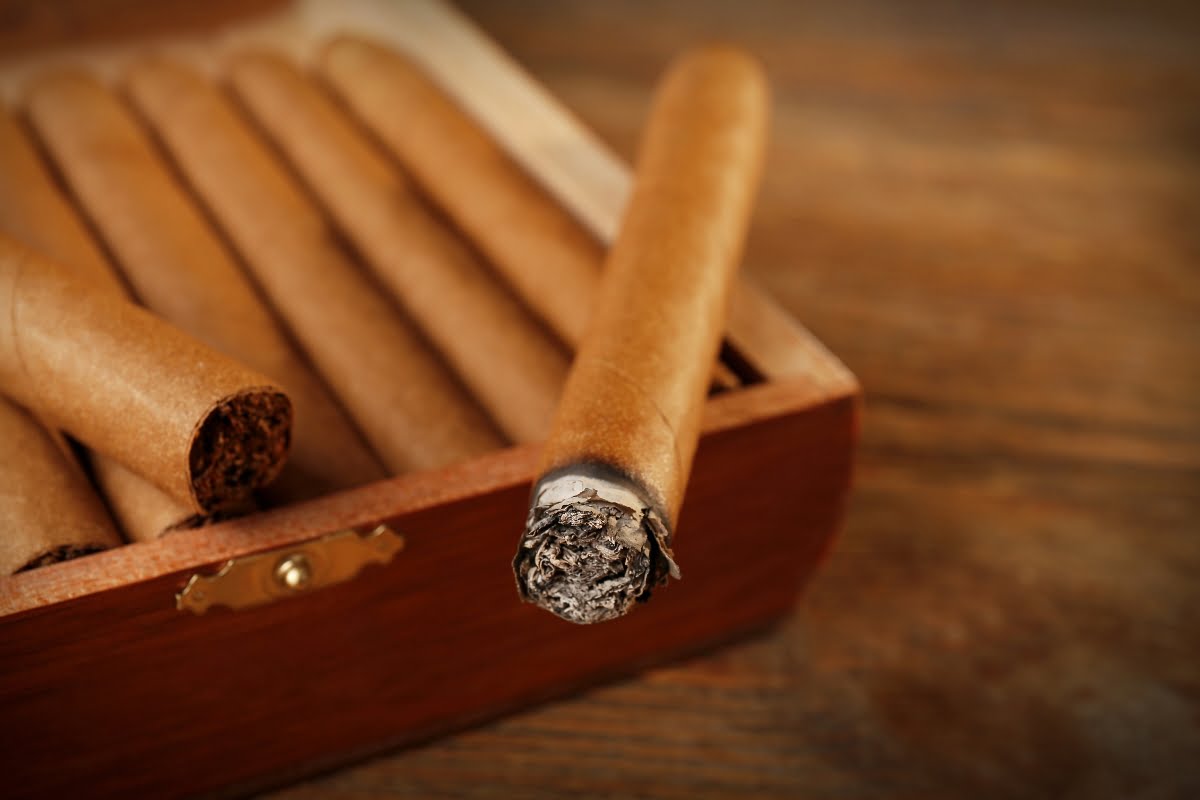 Close-up view of a partially burnt cigar resting on the edge of a wooden box filled with unlit cigars, creating a nostalgic scene for smoking memorabilia aficionados.