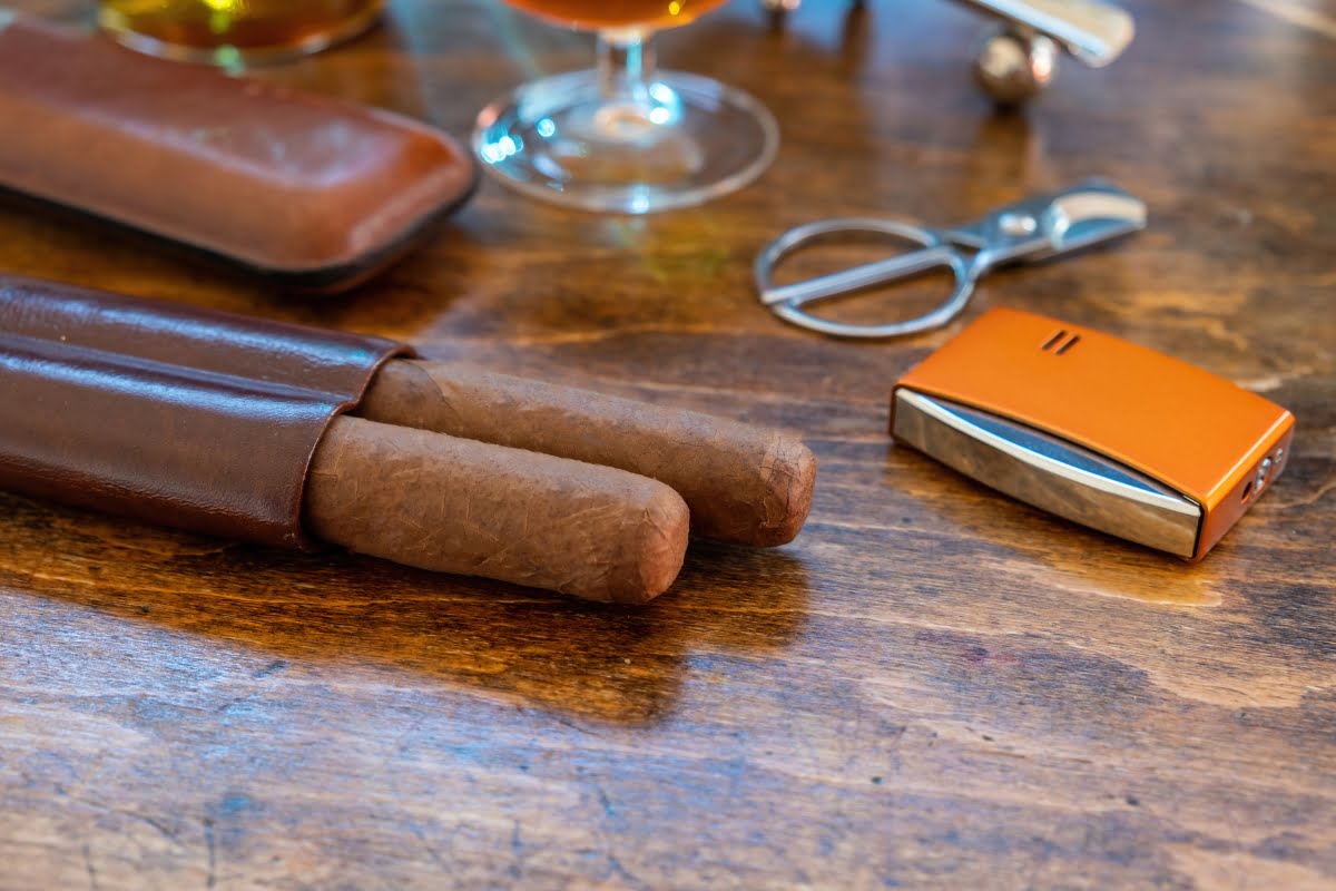 Two cigars in a leather case, a cigar cutter, and an orange lighter placed on a wooden surface—perfect smoking memorabilia for any aficionado.