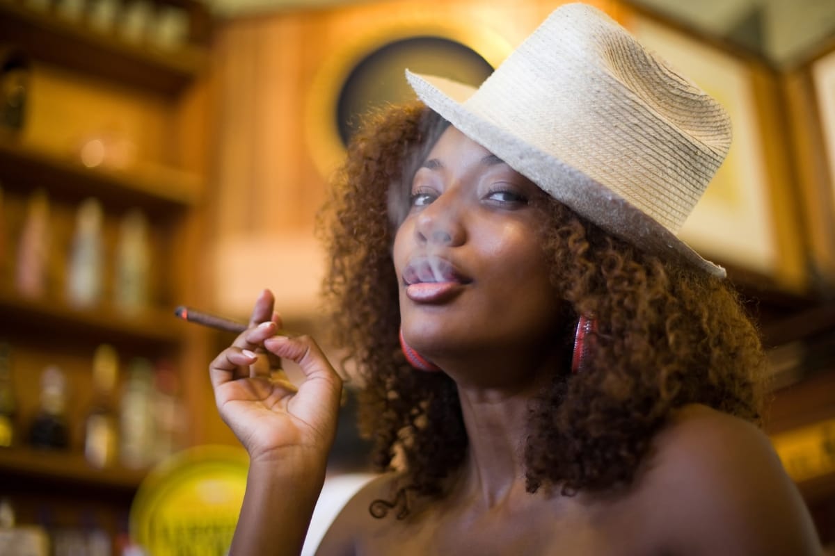 A person with curly hair wearing a white hat smokes a cigar indoors, surrounded by the rustic charm of wooden walls and shelves. The cozy setting evokes the timeless ambiance of Marietta Square.