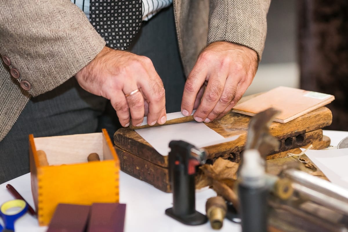 On a wooden table at Marietta Square, a person elegantly crafts cigars, using tools like a knife and small torch. Dressed in a suit jacket and patterned tie, they blend tradition with style in this charming locale.