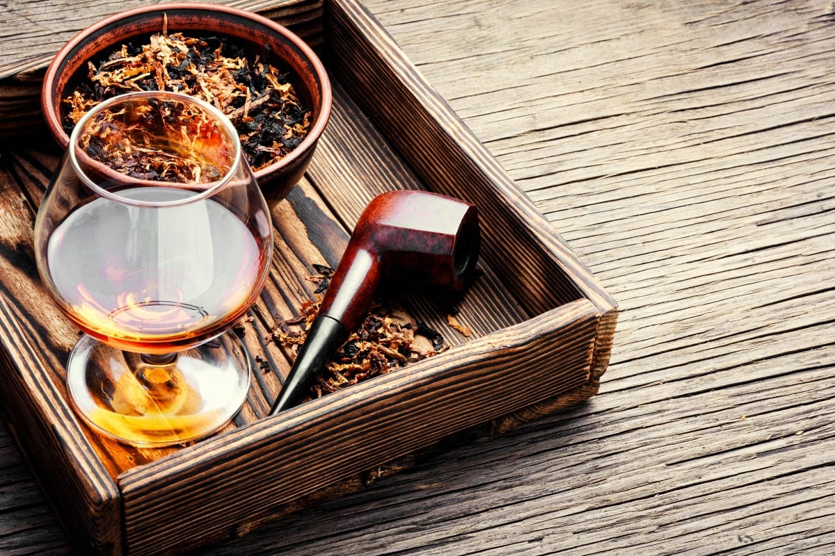 A wooden tray on the wooden surface of Marietta Square holds a glass of amber liquid, a tobacco pipe, and a bowl of dried tobacco.