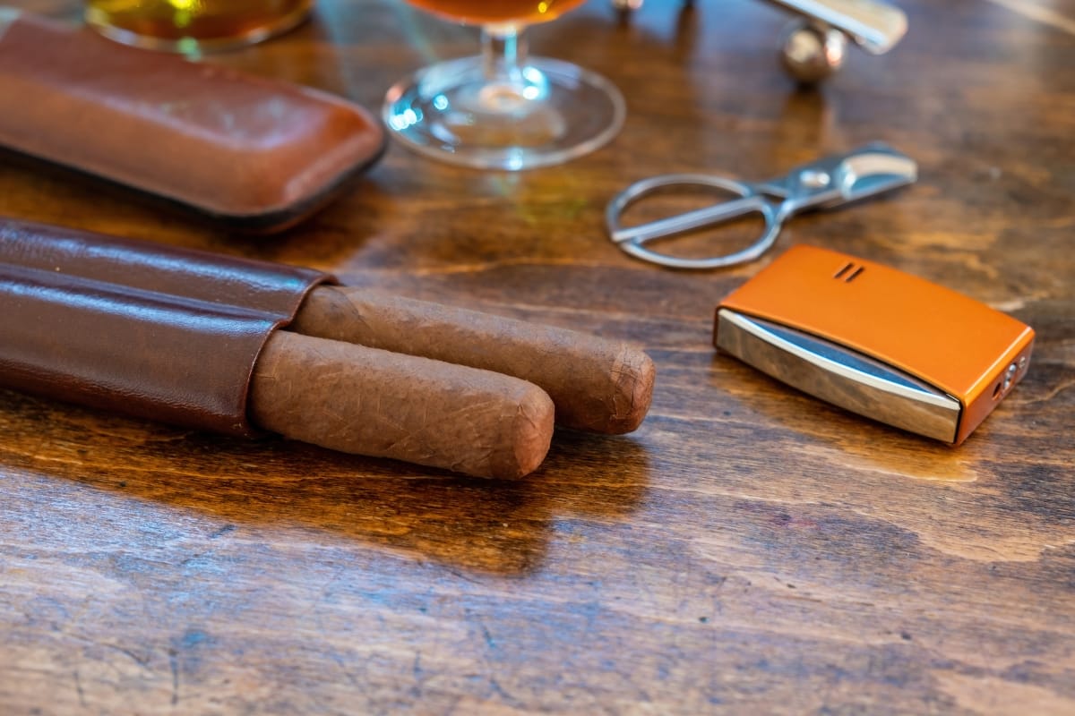 Two cigars in a leather case rest on a wooden table at Marietta Square, accompanied by a cigar cutter, an orange lighter, and a glass of amber liquid.