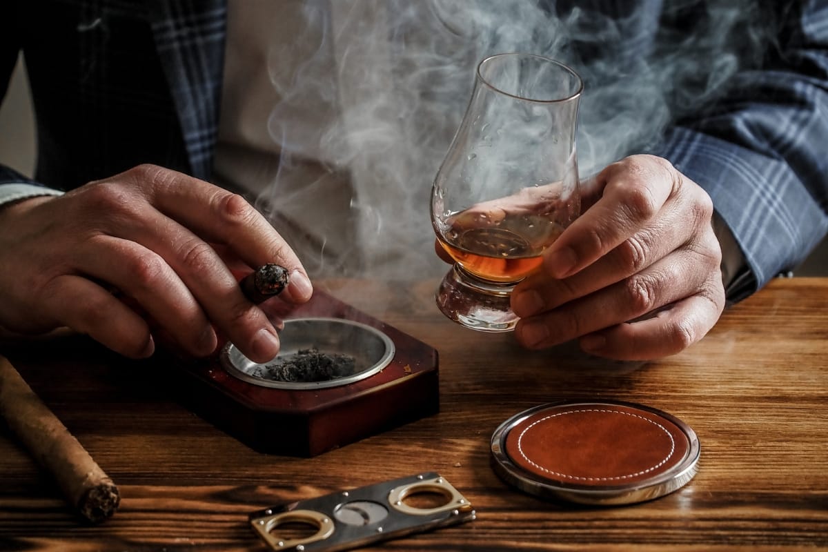 A person lounges at Marietta Square, holding a glass of whiskey in one hand and a lit cigar in the other, with an ashtray and cigar cutter resting on the wooden table beside them.