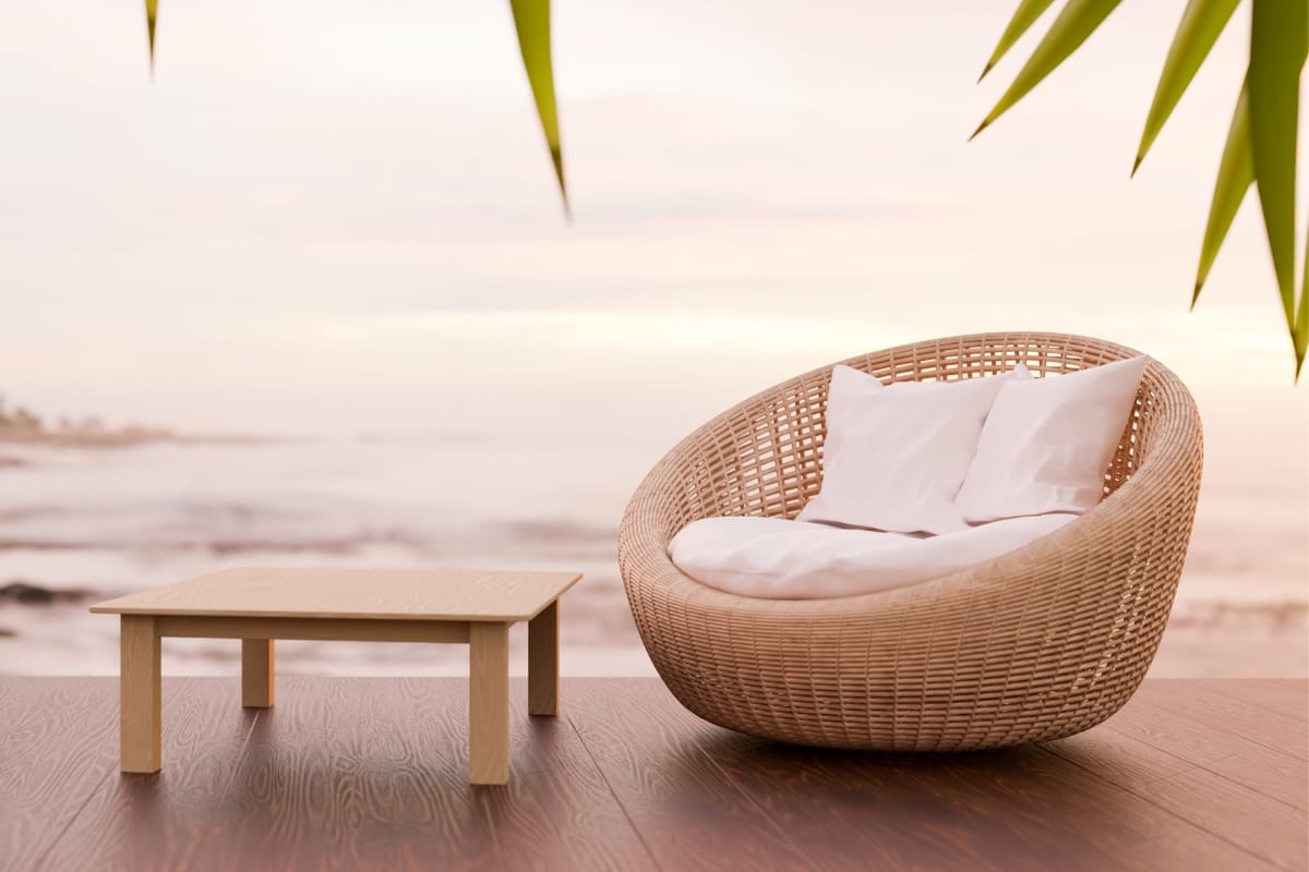 A rattan chair with white cushions sits next to a wooden table on a deck overlooking a calm ocean, creating an inviting outdoor smoking lounge, complemented by green leaves hanging above.